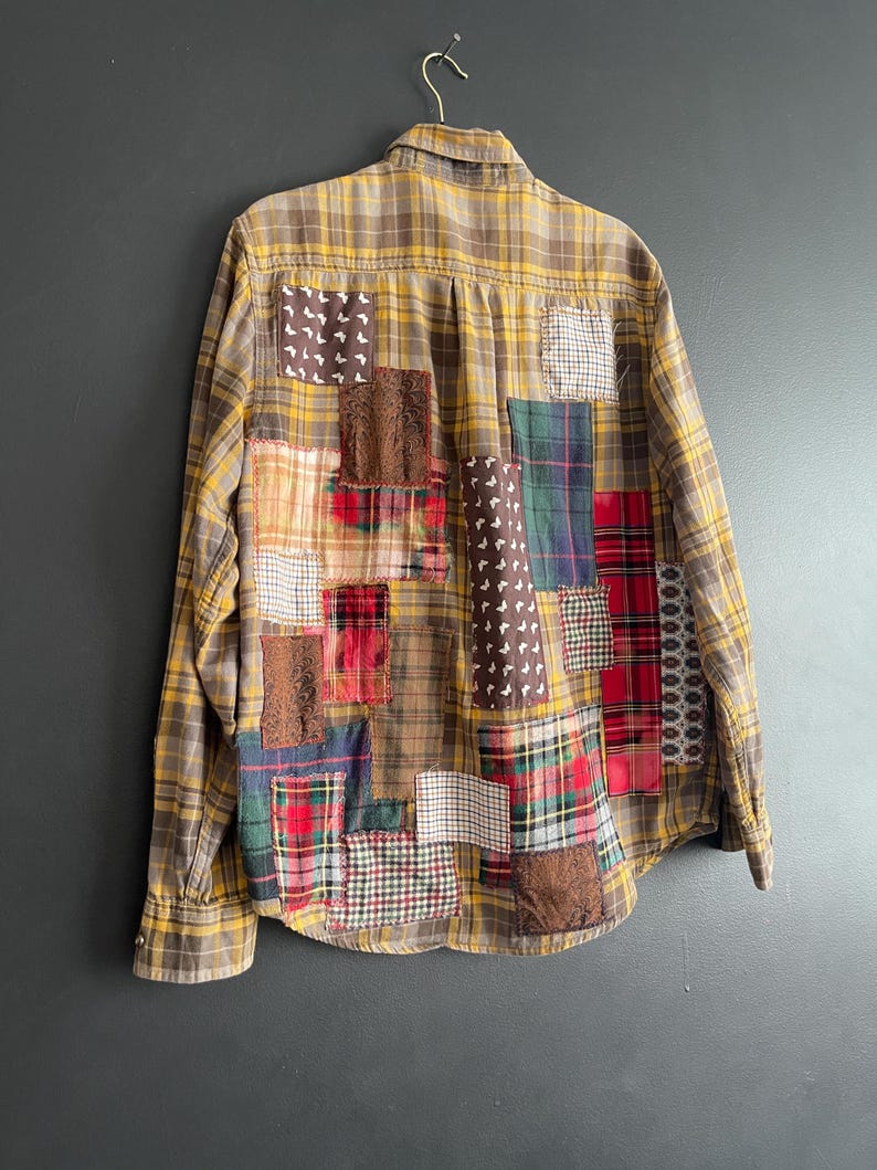 Op de afbeelding: Een button-down shirt met lange mouwen en een geel-bruin geruit patroon. De achterkant van het shirt is versierd met verschillende patches van verschillende stoffen, waaronder ruitjes, bloemen en textuurmaterialen. Het shirt hangt aan een metalen hanger.