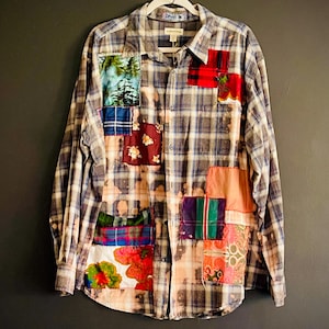 Op de afbeelding: Een overhemd met lange mouwen en knoopsluiting in een blauw-wit ruitpatroon met bleekvlekken. Het overhemd is versierd met diverse kleurrijke stoffen patches, waaronder bloemen-, bos- en ruitprints, die op de voorzijde zijn gestikt.