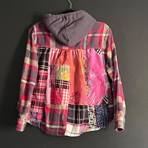 May include: A long-sleeve hooded shirt featuring a patchwork back panel composed of various fabric squares in shades of pink, red, and white. The upper portion and sleeves consist of a pink and grey plaid flannel fabric with an attached grey hood.