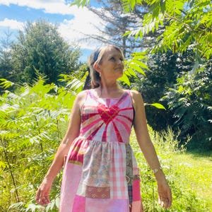 May include: A patchwork dress in shades of pink, red, and white, featuring a heart-shaped design on the bodice. The dress has a sleeveless design and a knee-length hemline. The background includes lush green foliage and a bright blue sky.