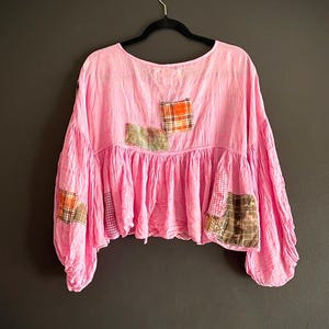 Op de afbeelding: Een roze top met lange mouwen en een ronde hals, versierd met patchwork details. De zoom is gerimpeld en de stoffen patches hebben verschillende ruitpatronen. De mouwen zijn wijd en gerimpeld bij de manchetten.