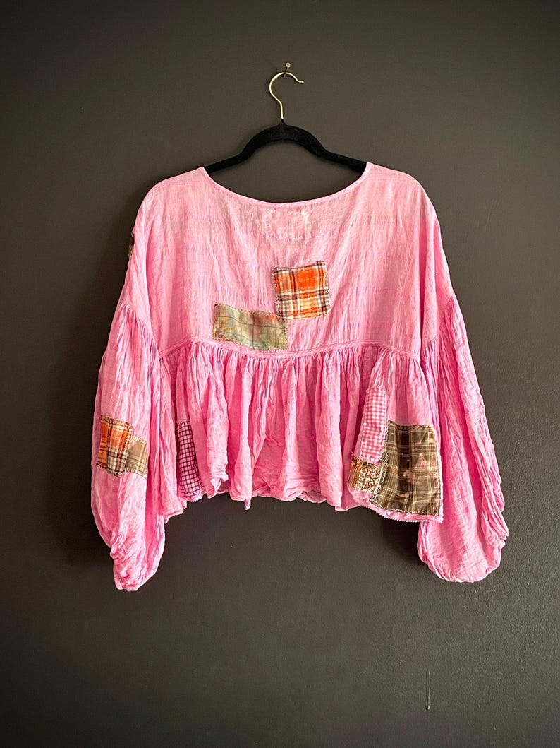 Op de afbeelding: Een roze top met lange mouwen en een ronde hals, versierd met patchwork details. De zoom is gerimpeld en de stoffen patches hebben verschillende ruitpatronen. De mouwen zijn wijd en gerimpeld bij de manchetten.