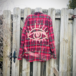 May include: Red and black plaid shirt with a bleached eye design on the back. The shirt is hanging on a weathered wooden fence. The eye design is in a cream color and has a sunburst design around it.