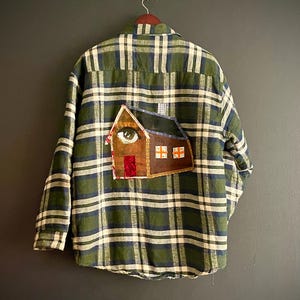 Könnte beinhalten: Grün, blau und weiß kariertes Flanellhemd mit einem hausförmigen Aufnäher auf dem Rücken. Der Aufnäher zeigt ein Auge, ein braunes Dach und eine rote Tür. Das Hemd hängt an einem Holzkleiderbügel.