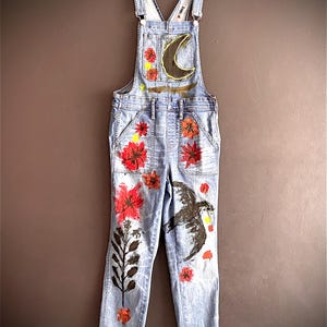 May include: Light blue denim overalls with hand-painted floral and celestial designs. Features a black crescent moon, red flowers, and a black bird. The overalls have a classic bib and brace design with frayed hems.