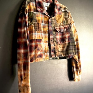 May include: A cropped long-sleeve button-down shirt constructed from various plaid flannel fabric patches in shades of mustard yellow, deep red, navy blue, and olive green. The garment features a front chest pocket, a collar, and raw hem edges.