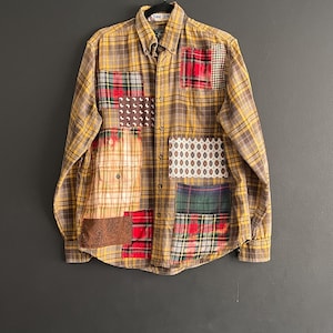 Op de afbeelding: Een button-down shirt met lange mouwen en een patchwork-ontwerp. Het shirt heeft een geel-bruin geruit patroon met verschillende stoffen patches in rood, groen en patronen. Het shirt hangt aan een zwarte hanger tegen een donkergrijze achtergrond.