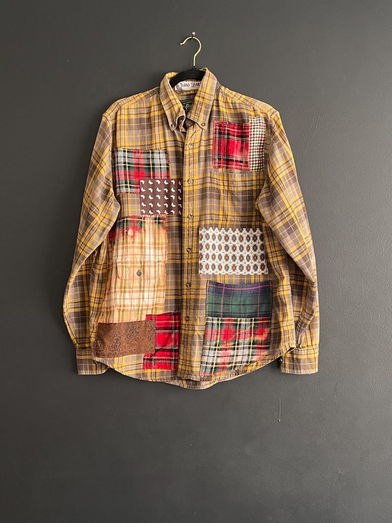 Op de afbeelding: Een button-down shirt met lange mouwen en een patchwork-ontwerp. Het shirt heeft een geel-bruin geruit patroon met verschillende stoffen patches in rood, groen en patronen. Het shirt hangt aan een zwarte hanger tegen een donkergrijze achtergrond.