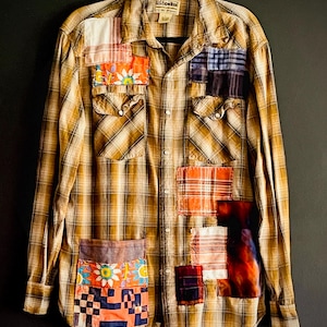 May include: A tan and brown plaid long-sleeve button-down shirt featuring various colorful fabric patches sewn onto the front. The patches include floral, checkered, and solid patterns in orange, blue, and white. The shirt has two chest pockets.