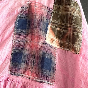Op de afbeelding: Close-up van een roze stoffen kledingstuk met geruite patches. De patches zijn met witte stiksels op de roze stof genaaid. E&eacute;n patch is een mix van blauw, roze en grijze ruit. De andere patch is bruin en tan geruit met een gebleekt effect.