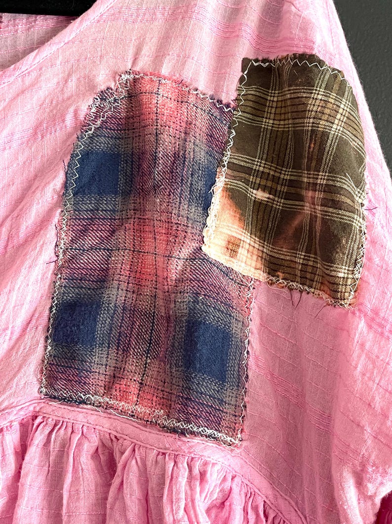 Op de afbeelding: Close-up van een roze stoffen kledingstuk met geruite patches. De patches zijn met witte stiksels op de roze stof genaaid. E&eacute;n patch is een mix van blauw, roze en grijze ruit. De andere patch is bruin en tan geruit met een gebleekt effect.