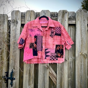 May include: Pink patchwork shirt with short sleeves, hanging on a black hanger. The shirt features a plaid pattern and various fabric patches in shades of pink, black, and floral designs. The shirt is hanging on a wooden fence.