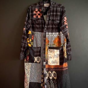 May include: A long, patchwork coat with a plaid upper section and a variety of patterned fabric panels. The coat features a button-down front, pockets, and accents of pink and orange. The coat is hanging on a black hanger.