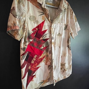 May include: Short-sleeved button-down shirt with a cream-colored floral pattern. A red and plaid lightning bolt design is sewn on the front. The shirt is hanging on a gray hanger.