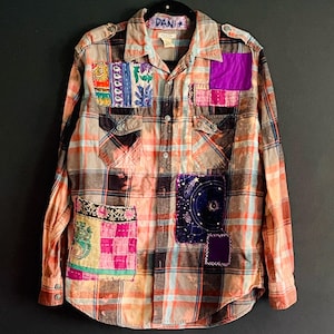 May include: A long-sleeve button-down shirt in a faded orange and blue plaid pattern, featuring various colorful fabric patches sewn onto the front, including a dark blue celestial print and a pink patterned square. The label reads DANI.