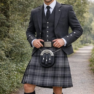 May include: A formal grey tweed suit with a matching waistcoat, white dress shirt, and black tie. The outfit includes a traditional Scottish kilt in a grey and black plaid pattern, a sporran, and black knee socks. The man is standing on a path.