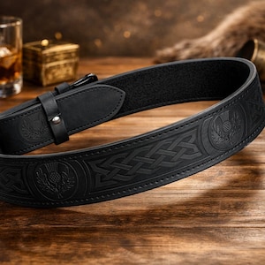 Puede incluir: Un cintur&oacute;n de cuero negro con dise&ntilde;os en relieve de nudos celtas y cardos. El cintur&oacute;n tiene un cierre de hebilla y se muestra sobre una superficie de madera. Un accesorio tradicional.