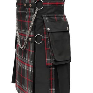 Może przedstawiać: Czarny kilt z czerwono-szarymi akcentami w kratę. Posiada łańcuch, metalowe pierścienie i kieszenie. Kilt ma plisowany design i strukturalny pas z srebrnymi elementami.