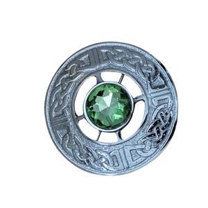 May include: A silver-toned brooch with a Celtic knot design around a central, faceted, green gemstone. The brooch is circular with intricate detailing, and the gemstone is a vibrant green color.