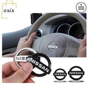 Peut inclure: Remplacement d'emblèmes de volant Nissan argentés et noirs. L'emblème est présenté dans une main, avec le volant en arrière-plan. Le mot "NISSAN" est imprimé sur l'emblème. L'image montre également le volant avec le logo Nissan.