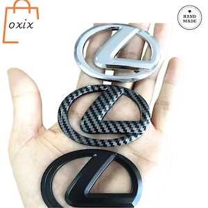 Peut inclure: Trois emblèmes de voiture en argent, fibre de carbone et noir. Les emblèmes présentent le logo Lexus. L'emblème supérieur est argenté brillant, celui du milieu a un motif en fibre de carbone et celui du bas est noir mat. Le mot "oxix" est dans le coin supérieur gauche.