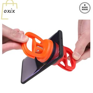 Puede incluir: Una herramienta de ventosa naranja y negra se utiliza para levantar un teléfono inteligente negro. La herramienta tiene un mango rojo y una ventosa negra. El teléfono está sostenido por dos manos. La imagen también incluye el texto "oxix" y "Hand Made".