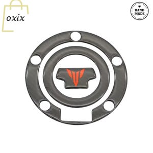 May include: A dark gray motorcycle fuel tank pad with a red and orange logo. The pad is circular with a central design and several mounting holes. The word "oxix" is in the upper left corner, and a "Hand Made" logo is in the upper right.