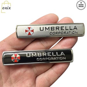 May include: Two rectangular metal badges with the text "UMBRELLA CORPORATION". One badge is silver with a red, white, and black logo. The other is black with the same logo. The badges are handmade.