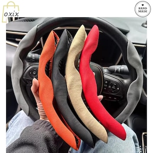 Puede incluir: Una selección de fundas de volante coloridas en naranja, negro, beige y rojo, cada una con un diseño perforado. Las fundas se sostienen frente al volante de un coche, mostrando su forma y textura. La imagen incluye una pegatina "Hand Made".