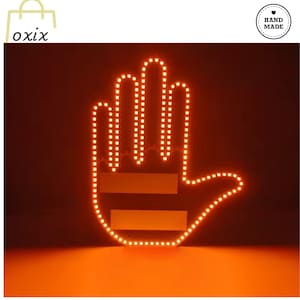 Puede incluir: Un letrero iluminado en forma de mano de color naranja con un borde de pequeñas luces. Dos rectángulos horizontales están colocados en el área de la palma. El letrero está contra un fondo oscuro. La palabra "oxix" está en la esquina superior izquierda y un logotipo de "Hand Made" está en la parte superior derecha.