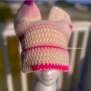 May include: A handmade crochet hat in cream and pink, featuring a cat ear design. The hat has a bright pink brim and accents, with the text "@BLESSEDLOOPS.CO" visible on the side. The hat is displayed on a mannequin head.