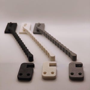 K&ouml;nnte beinhalten: Eine Auswahl an 3D-gedruckten Kunststoffkomponenten in Schwarz, Wei&szlig; und Grau. Die Komponenten umfassen Hebel mit geriffelten Griffen und passende quadratische Teile mit einem hakenartigen Design. Die Artikel sind auf einer wei&szlig;en Oberfl&auml;che angeordnet.