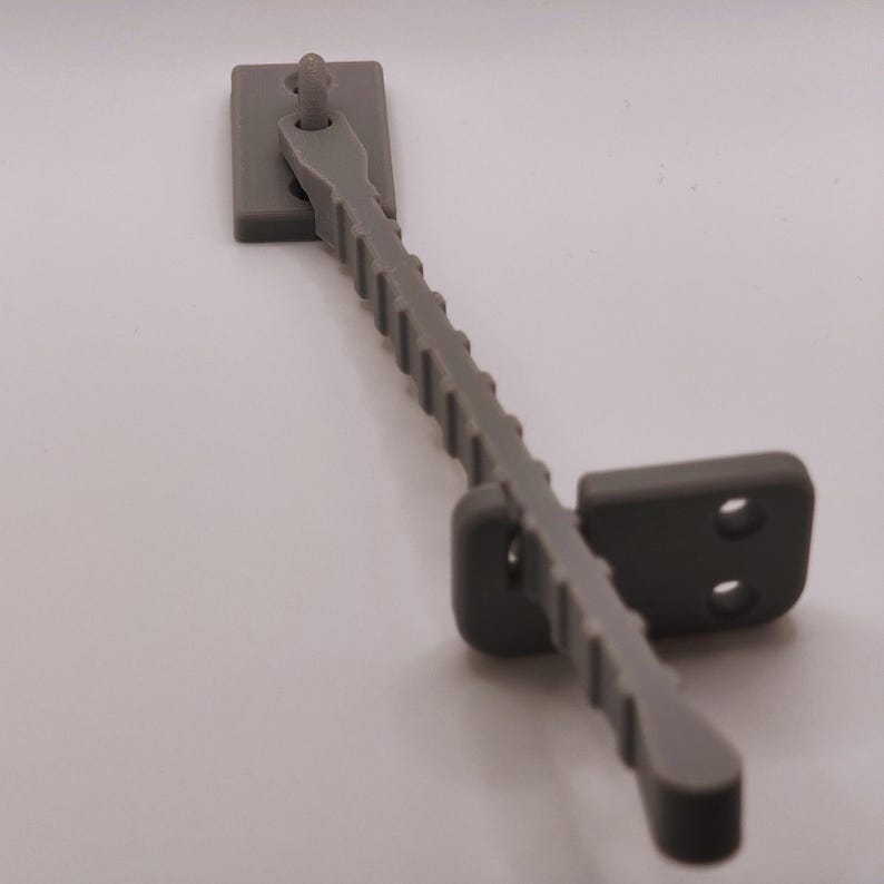 K&ouml;nnte beinhalten: Ein grauer Fensterriegel aus Kunststoff. Er hat eine rechteckige Basis, einen geriffelten Arm und ein zweites rechteckiges Teil mit zwei L&ouml;chern. Der Arm ist angelenkt. Dieser Riegel dient zur Sicherung eines Fensterfl&uuml;gels.