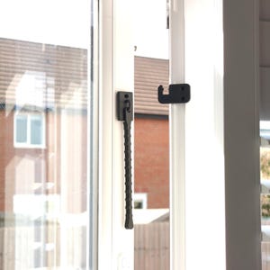 K&ouml;nnte beinhalten: Nahaufnahme eines wei&szlig;en Fensterrahmens mit einem schwarzen Fensterriegel und Haken. Der Riegel hat einen strukturierten Griff. Das Fenster spiegelt eine Ansicht eines Backsteingeb&auml;udes und eines Wohngebiets wider.