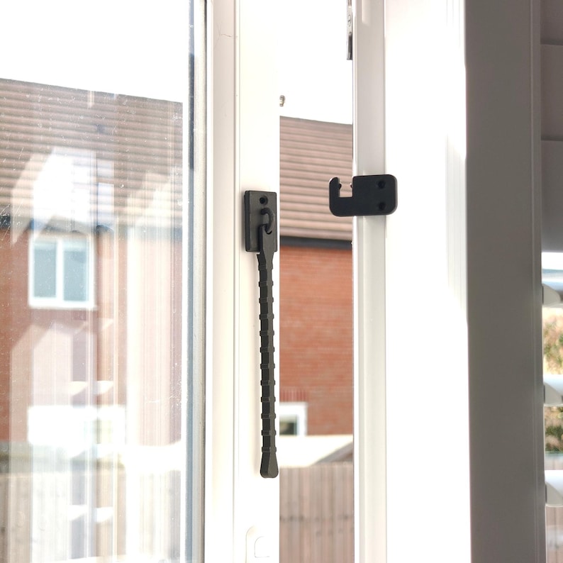 K&ouml;nnte beinhalten: Nahaufnahme eines wei&szlig;en Fensterrahmens mit einem schwarzen Fensterriegel und Haken. Der Riegel hat einen strukturierten Griff. Das Fenster spiegelt eine Ansicht eines Backsteingeb&auml;udes und eines Wohngebiets wider.