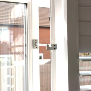 K&ouml;nnte beinhalten: Ein wei&szlig;er Fenstersicherheitsverschluss, der an einem wei&szlig;en Fensterrahmen angebracht ist. Der Verschluss besteht aus grauem Kunststoff und hat ein flexibles, geripptes Band, das zwei Montageplatten verbindet. Das Fenster ist leicht ge&ouml;ffnet und gibt den Blick auf ein Wohngebiet frei.