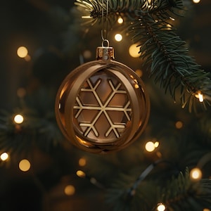 Puede incluir: Un adorno navide&ntilde;o dorado con un dise&ntilde;o de copo de nieve. El adorno cuelga de una rama verde de un &aacute;rbol de Navidad. El fondo est&aacute; borroso con peque&ntilde;as luces desenfocadas.