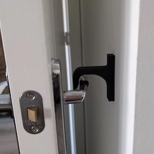 May include: A black door handle helper attached to a white door. The handle helper is a T-shaped design, made of a solid black material. The door handle is silver.