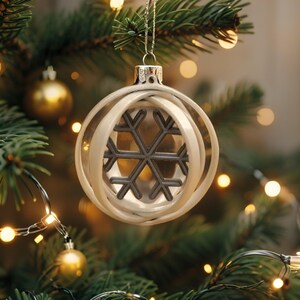 Puede incluir: Una bola de Navidad de cristal transparente con un dise&ntilde;o de copo de nieve. El adorno tiene una tapa plateada y est&aacute; suspendido de un hilo plateado. El copo de nieve es de color gris oscuro y est&aacute; rodeado por un dise&ntilde;o blanco en forma de cinta. El adorno cuelga de un &aacute;rbol de Navidad.