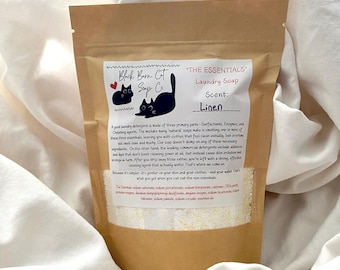 May include: A brown paper bag of "Linen" scented laundry soap. The label features a black cat logo and the text "Black Born Cat Soap Co." The bag is filled with white powder and rests on a white fabric surface.