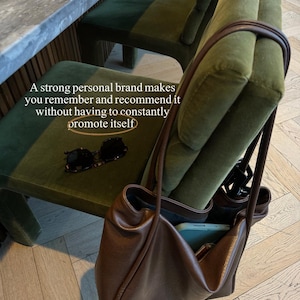 May include: A brown leather tote bag with a soft, slouchy design, resting on a green velvet chair. The bag contains a phone. Sunglasses are on the chair. The text reads: "A strong personal brand makes you remember and recommend it without having to constantly promote itself."