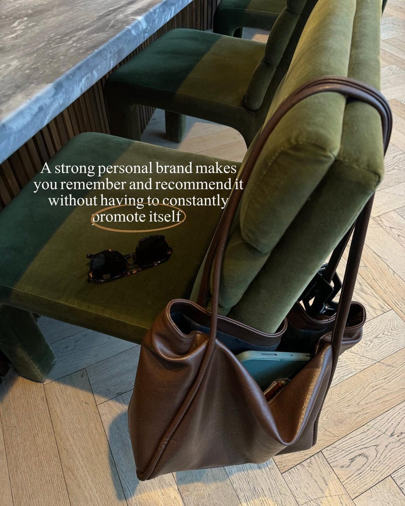 May include: A brown leather tote bag with a soft, slouchy design, resting on a green velvet chair. The bag contains a phone. Sunglasses are on the chair. The text reads: "A strong personal brand makes you remember and recommend it without having to constantly promote itself."