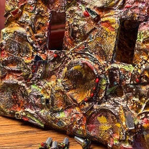 May include: A close-up of a colorful, textured light switch cover with a mosaic-like design. The cover is made of various materials, including metal, glass, and paint. Four small screws are visible in the foreground.