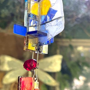 May include: A colorful, abstract glass art piece with a mix of blue, yellow, red, and clear glass elements. The design includes geometric shapes and a faceted red bead. The piece is suspended by a silver-colored wire.