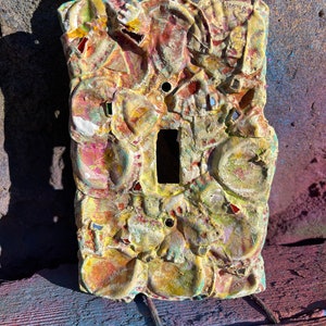May include: A light switch cover made of colorful mosaic tiles. The tiles are arranged in a random pattern and are mostly shades of yellow, orange, red, and green. The switch cover is rectangular and has a single light switch in the center.