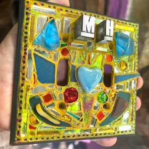 May include: A mosaic light switch cover with a black border and a yellow background. The cover is decorated with colorful glass pieces, including blue, red, and mirrored fragments. The letters "MH" are displayed on the top.