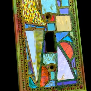 May include: A rectangular light switch cover plate decorated with a mosaic of multi-colored glass pieces in shades of blue, orange, yellow, and purple. The frame is painted in a textured lime green with small raised dots along the border.