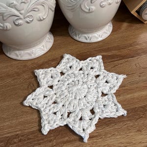 May include: A white, star-shaped crochet coaster. The coaster is made of thick, textured yarn and features a detailed openwork design. The coaster is placed on a wooden surface, with two white decorative vases in the background.