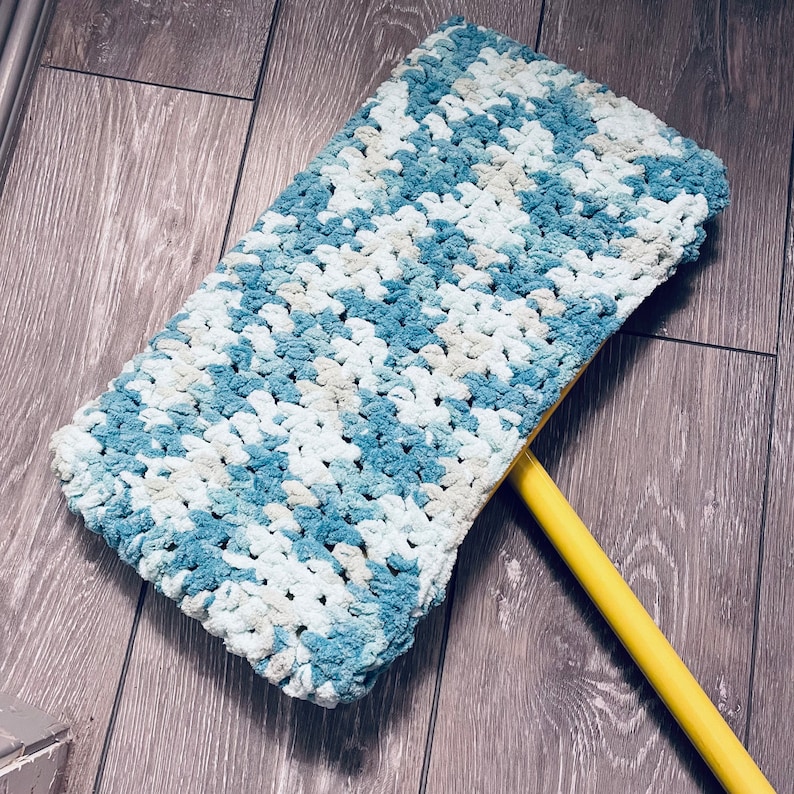 May include: A blue and white crocheted mop head attached to a yellow handle. The mop head is made of soft, fluffy yarn and has a textured, crocheted design.