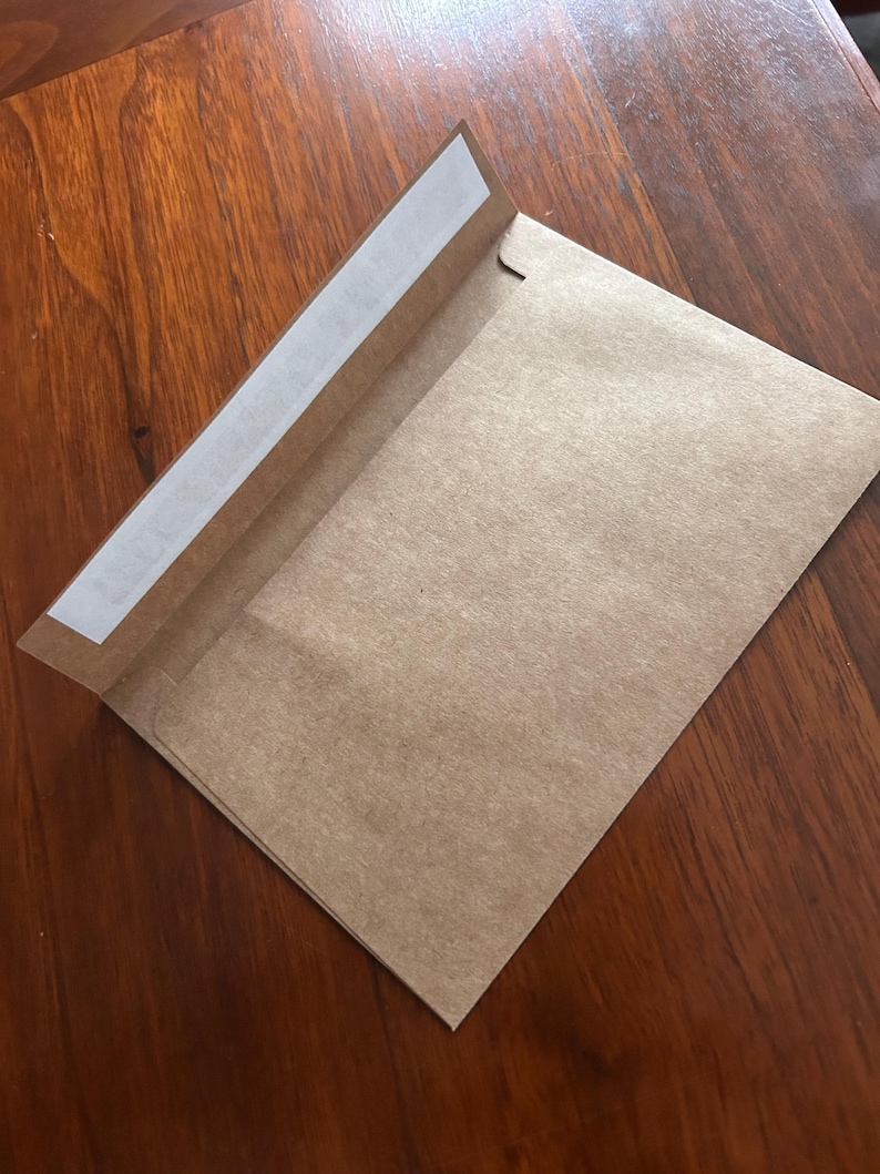 Może przedstawiać: Brązowa koperta papierowa z białym paskiem samoprzylepnym na klapce. Koperta jest otwarta, odsłaniając wnętrze. Tło stanowi drewniana powierzchnia.